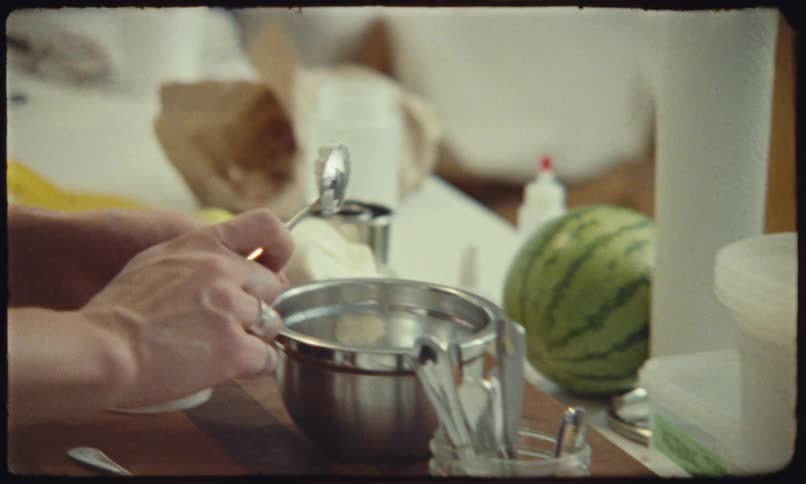 A person wearing a dark apron works at a kitchen counter carefully handling a small white object that appears to be a round of cheese or a pastry component