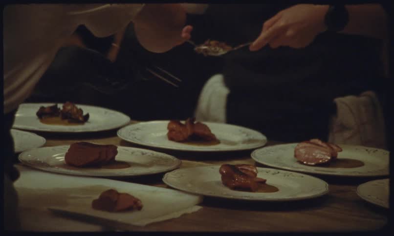 In a professional kitchen setting chefs wearing black gloves and white shirts carefully plate slices of seared meatthey use spoons and tweezers to add sauces and garnishes to several plates lined up on a wooden counter