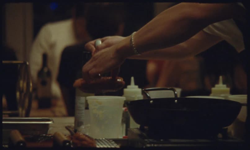 A person prepares food over a pan in a dimly lit kitchen environment with customers around eating and drinking