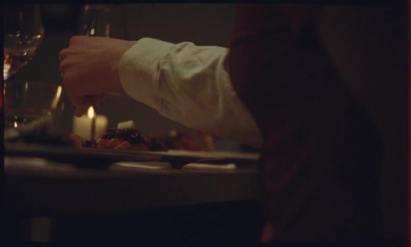 A close up shot shows a persons hands using a fork to eat a meal during a candlelit dinner in a dimly lit setting