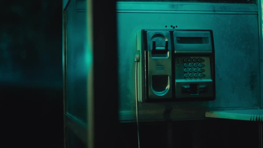A weathered silver payphone hangs inside a dimly lit glass booth bathed in a moody teal and blue light the camera slowly pushes in on the keypad and receiver capturing a lonely cinematic atmosphere at night