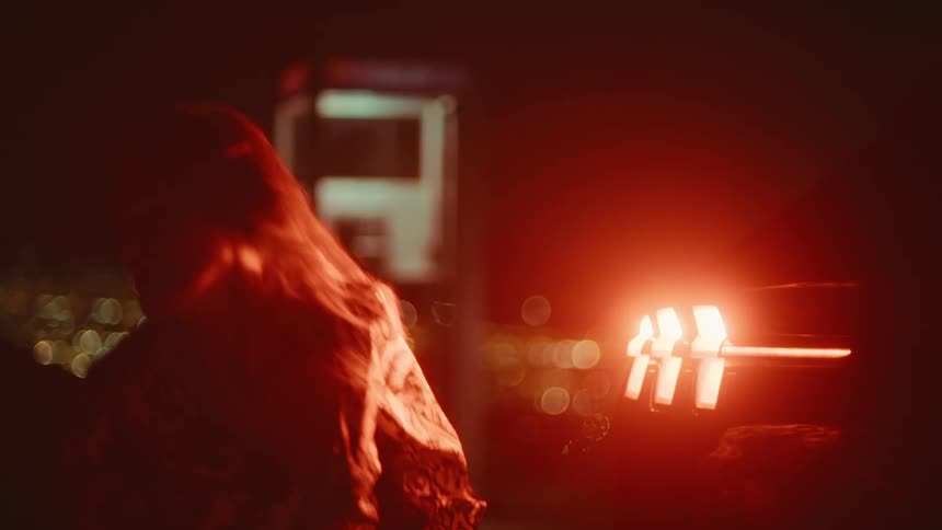 Woman crouches next to the glowing tail lights of a vehicle then she stands and walks away
