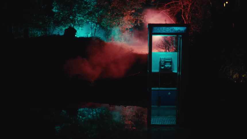 In a dark wooded area an eerie blue light illuminates an isolated telephone booth until mysterious red smoke drifts by and a bright warm light flashes inside