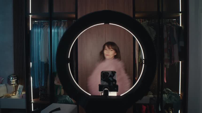 Young adult woman wearing a pink fluffy jacket dancing in front of a smartphone in a bedroom indoors during day time
