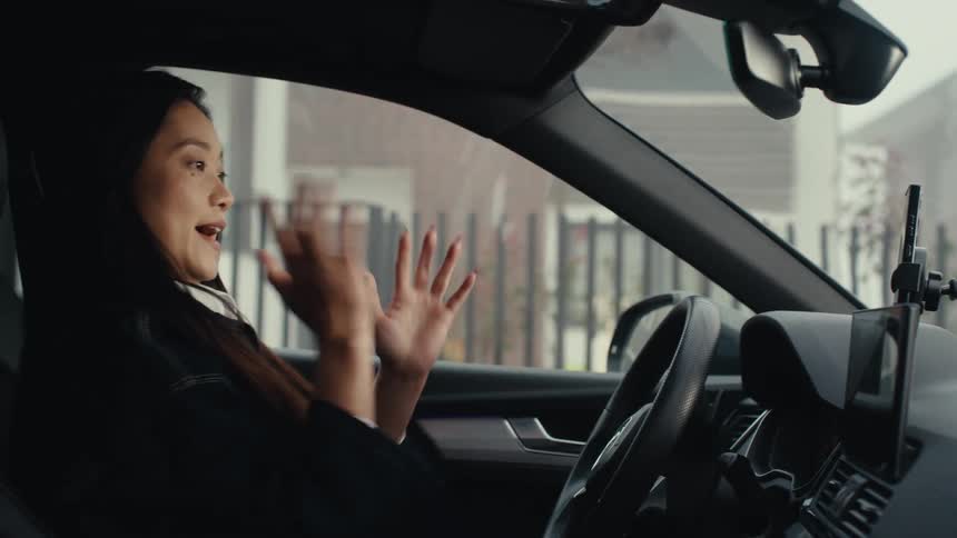 A woman sits in the drivers seat of a car and attaches her smartphone to a dashboard mount before an older man approaches her window to start a conversation while she keeps her hands on the steering wheel