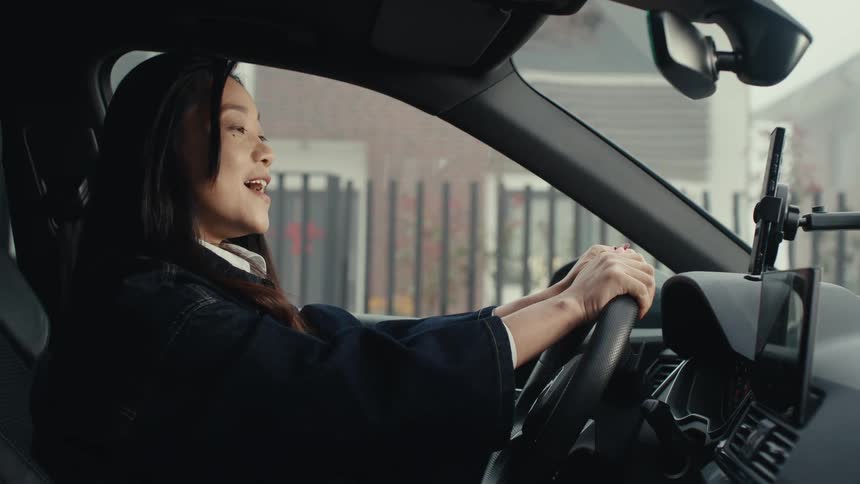 A woman sitting in the drivers seat of a car interacts with a mobile phone mounted on the dashboard and then speaks with an older man who approaches her window from outside