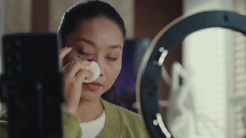 Young adult woman applying makeup with a sponge and talking in front of a smartphone indoors during day time