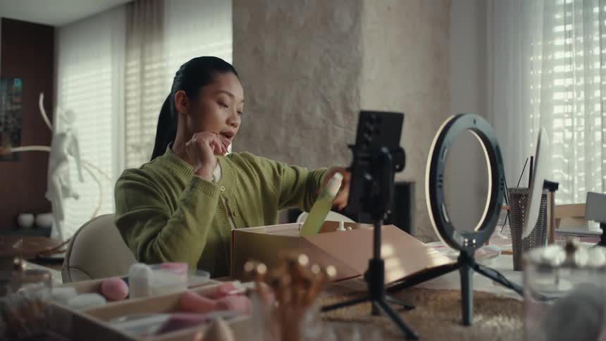 An asian woman in a green sweater sits at a table filming a skincare unboxing video using a smartphone and ring lightshe holds various bottles up to the camera and gestures with her hands while explaining the products to her audience