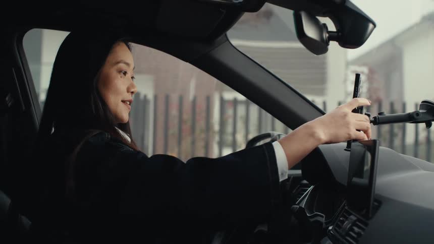 A female sits inside a vehicle mounting a smart phone to the dashboard and speaks into the phone in a suburb area