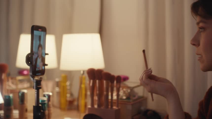 Woman records a makeup tutorial in front of a mirror with a phone on a tripod and table lighting