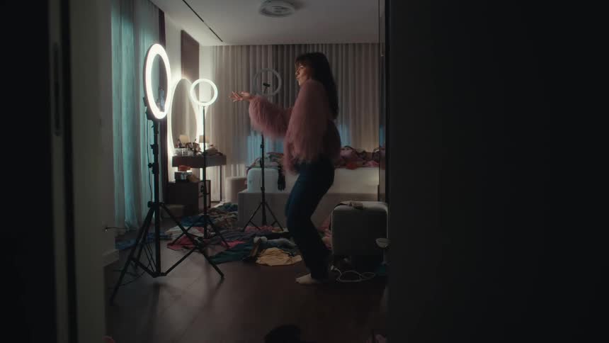 A woman in a purple faux fur jacket and blue jeans dances in a dimly lit cluttered messy bedroom looking forward with a joyful and energetic mood