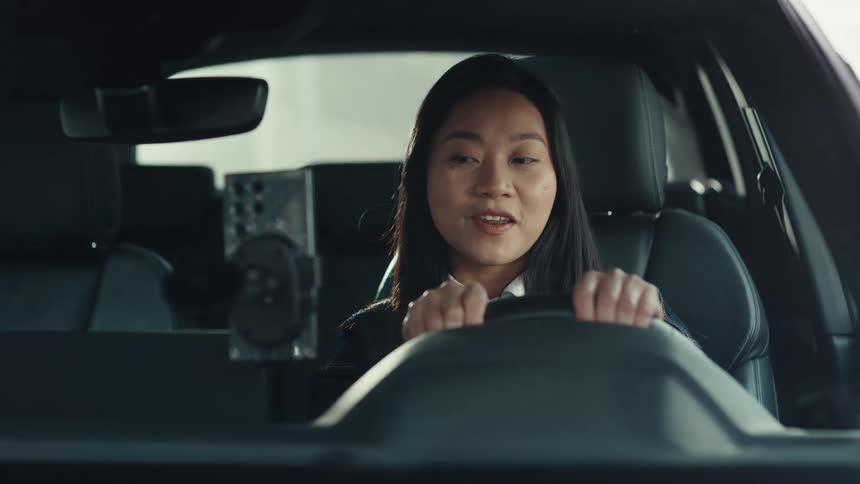 A woman with long dark hair sits in the drivers seat of a car and speaks to a mobile phone mounted on the dashboard