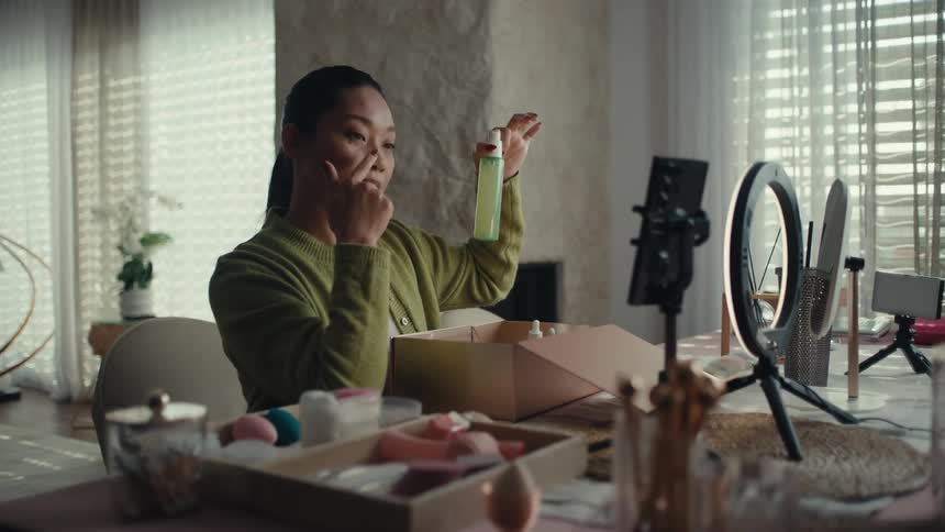 A woman sits at a table in front of a ring light and smartphone filming herself unboxing and demonstrating various skincare products from a package while speaking directly to the camera in a bright modern living room setting