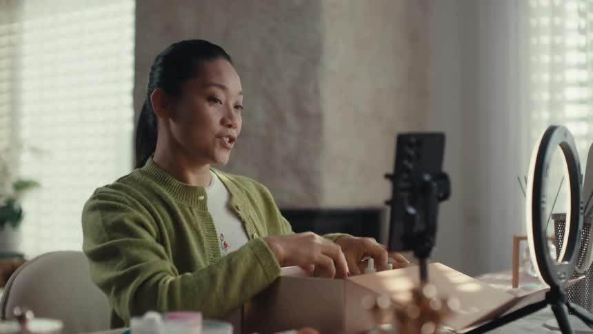 Young adult woman sitting in front of a table recoding a video on a smartphone while she applies makeup and shows products from a cardboard box indoors during day time