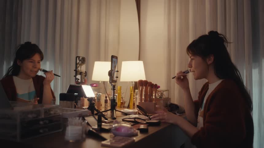 Woman applies makeup at a vanity while recording a video with a phone on a tripod