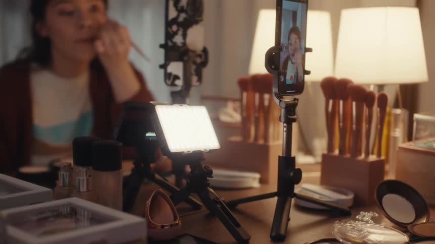 Woman applies makeup while recording a tutorial with a phone on a tripod in front of a mirror