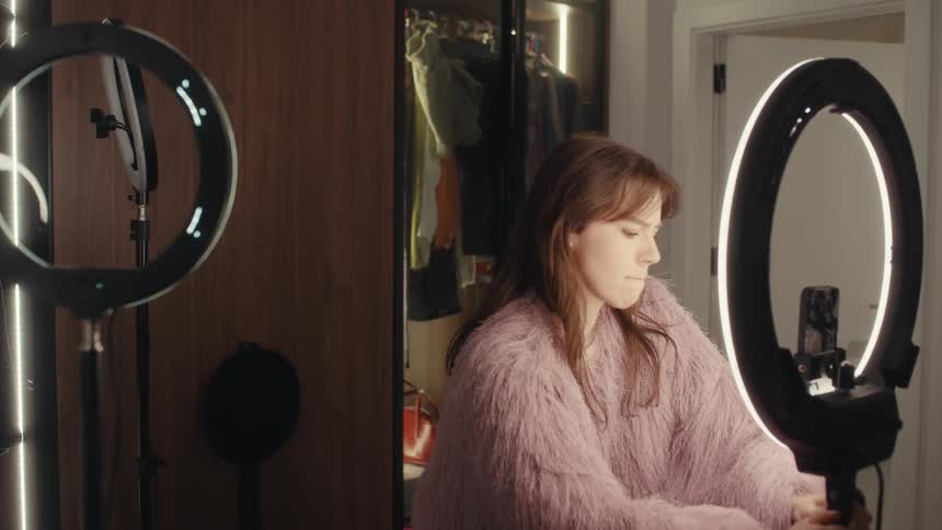 A woman in a shaggy pink jacket positions two ring lights in a room and she adjusts a phone on a tripod checks her appearance and begins dancing and spinning for the camera while a lit closet and a white door remain in the background
