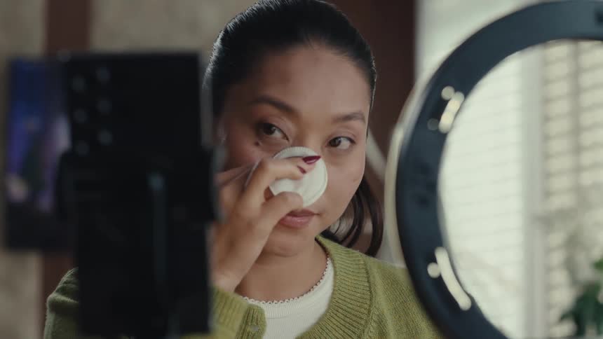 Young adult woman applying makeup in front of a smartphone indoors during day time