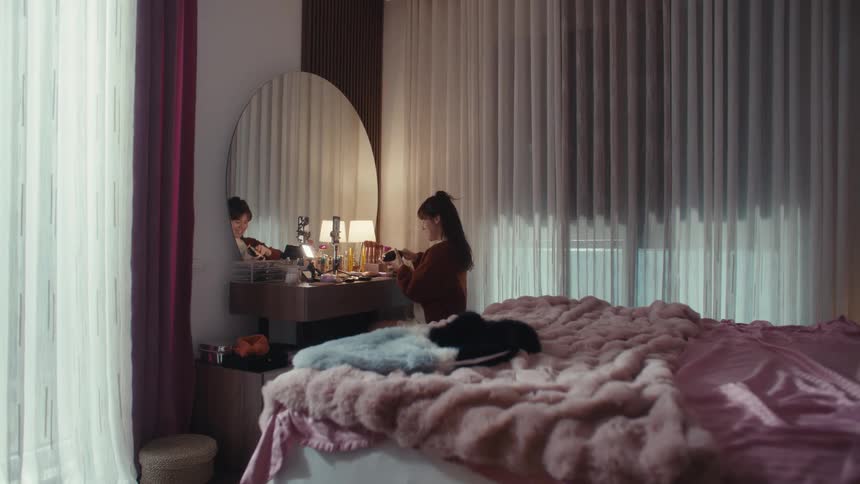 Woman applies makeup at a vanity while recording a video in a softly lit bedroom