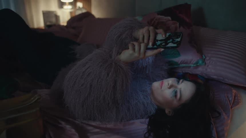 Teenage girl walks across the room and falls onto the bed as she looks at her phone in her hands