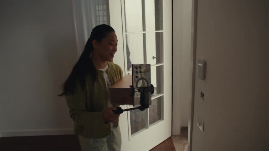 Young adult woman standing up from a chair while recording a video on a smartphone then walking to a door to pick up a cardboard box before going back to sit on the chair indoors during day time