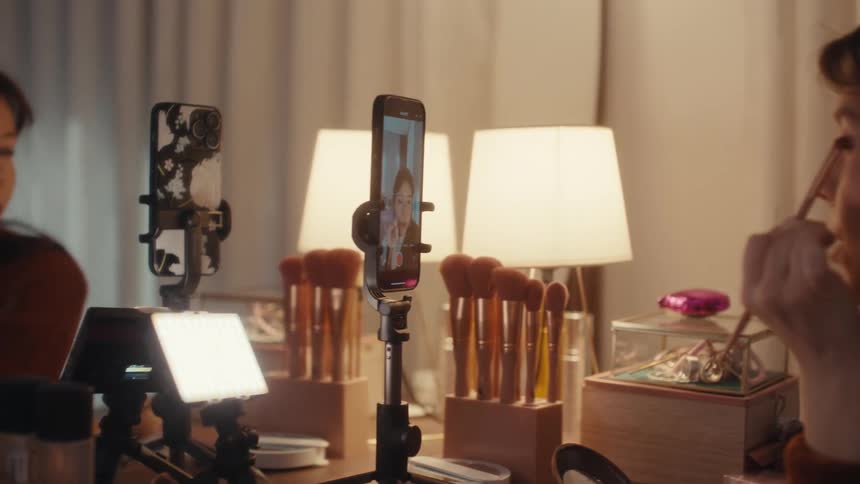 A young woman applies eyeshadow while recording a makeup tutorial using a smartphone mounted on a tripodshe carefully blends the product onto her eyelid while looking into the camera and speaking to her audience