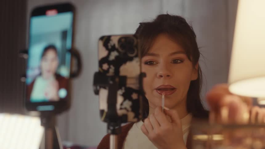 Woman applies lipstick while recording a tutorial with a phone on a tripod indoors