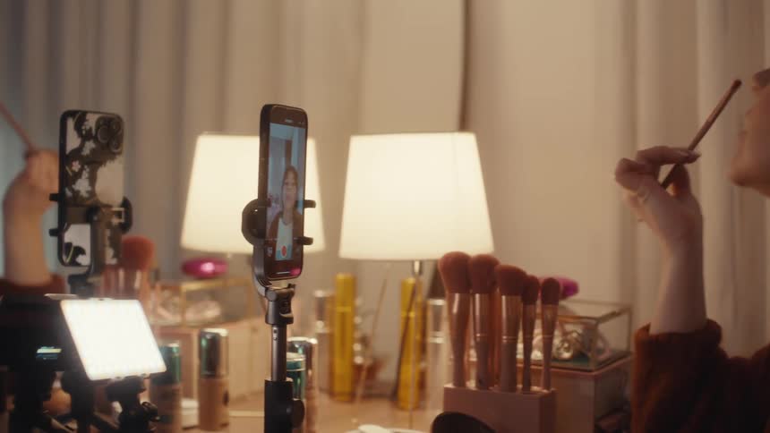 A young woman with brown hair and bangs sits at a desk covered in cosmetics and recording equipment applying eyeshadow with a brush while filming herself with a mobile phone