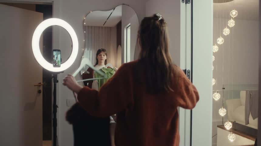 A woman in a brown sweater looks into a wavy mirror holding up different clothing pieces while a ring light holds a smartphone