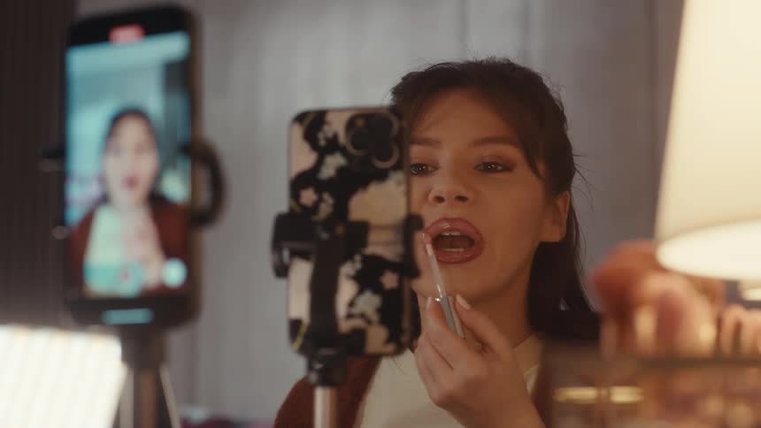 Woman applies lipstick while recording a tutorial with a phone on a tripod in front of a mirror
