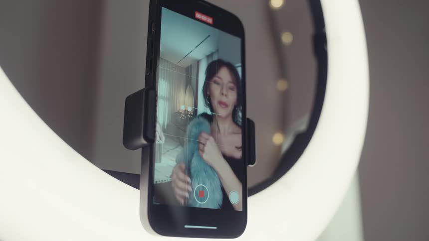 Woman appears on a phone screen mounted in a ring light while recording a video indoors