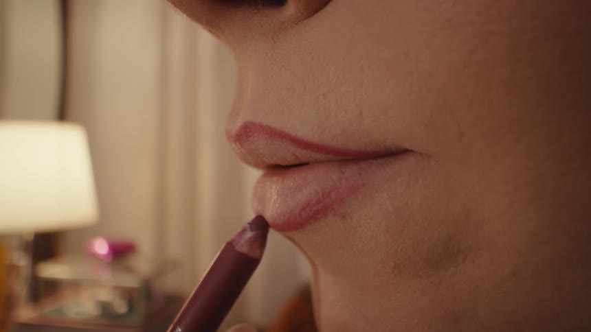 A woman carefully applies a dark red lipliner to the contours of her upper and lower lips in a closeup shot using a steady hand to define her mouth while sitting in a dimly lit room with a lamp glowing softly in the background