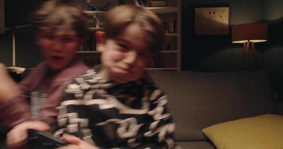 Two children sitting on a couch hold video game controllers in a lit living room