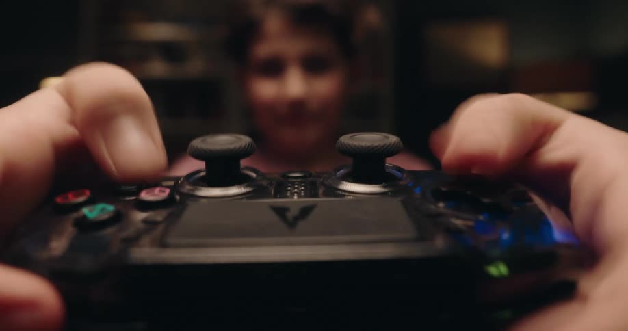 A closeup shot shows a persons hands holding and using a black gaming controller while a persons face is visible in the blurred background