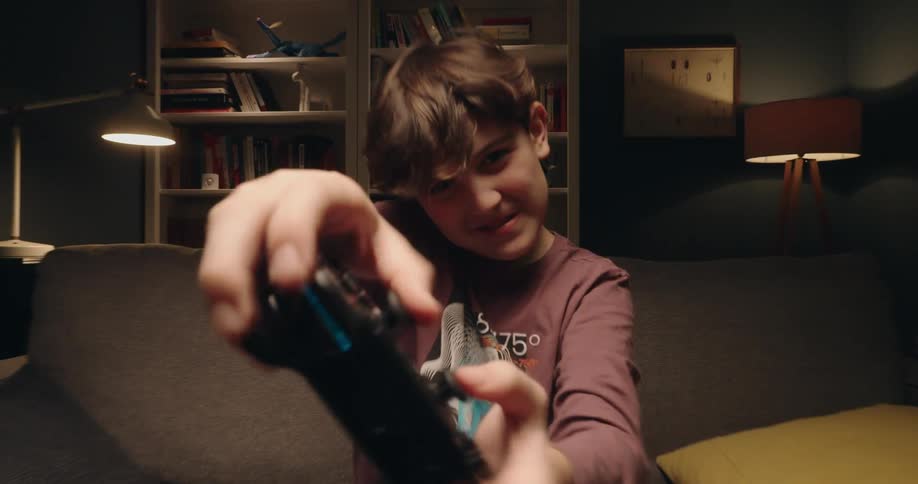 Boy holds a video game controller pointed forward while sitting on a couch
