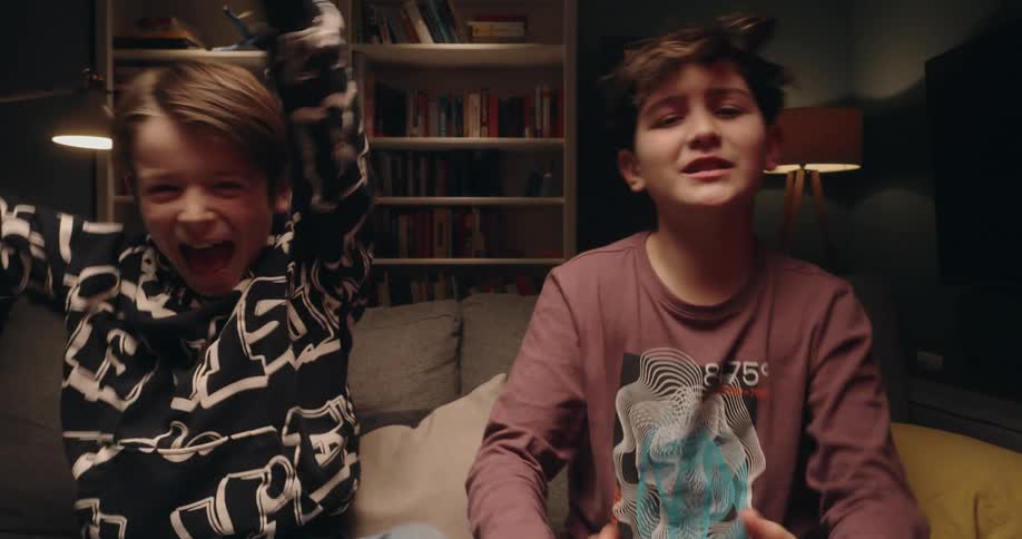Two young boys playing video games with controller while sitting on a couch and one of them smiling and cheering whilte the other throwing his body down in a sad manner indoors during night time