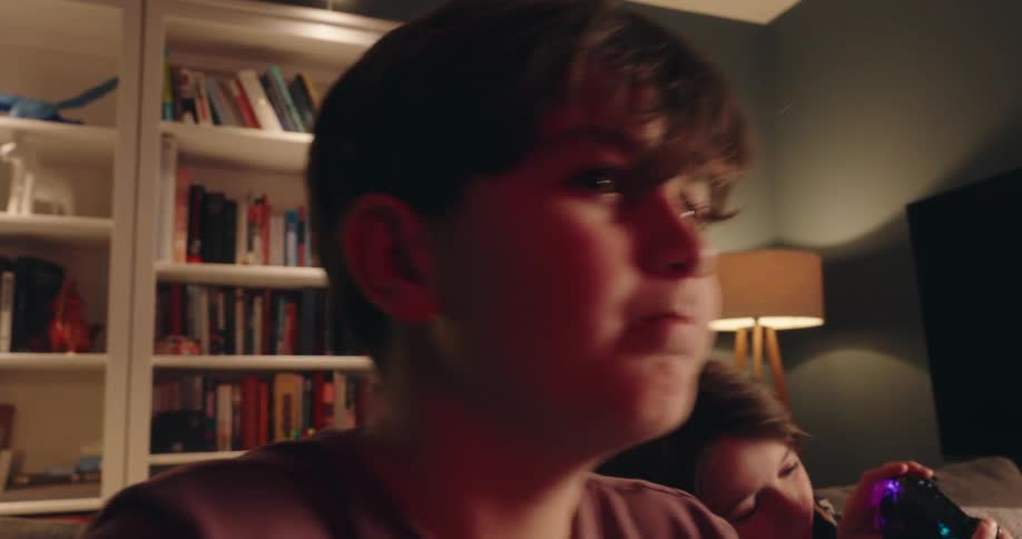 A young boy stares intently at a screen with a colorful glow reflecting on his face while gaming in a dimly lit living room as another child sits partially visible behind him holding a controller near a bookshelf and a lamp