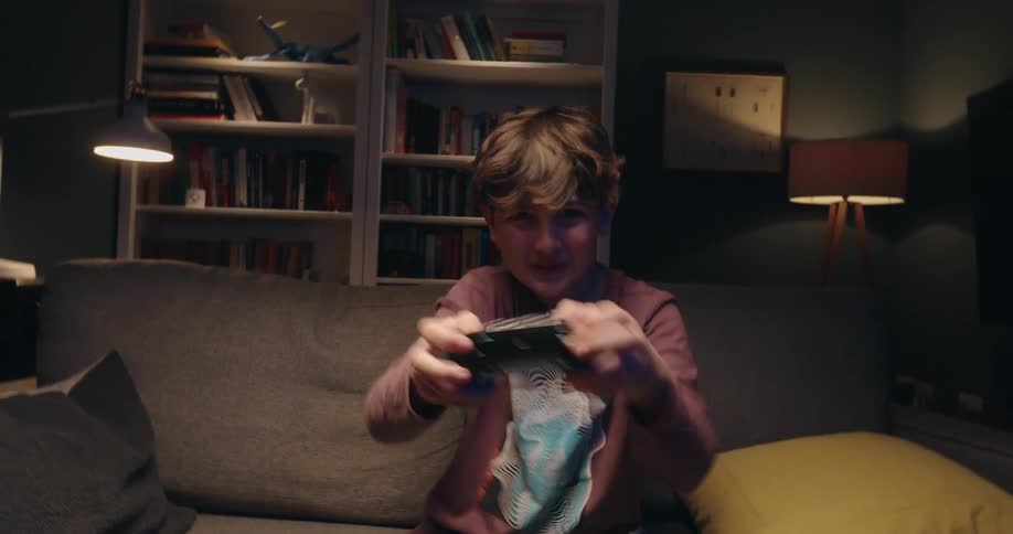 A young boy sits on a couch in the living room at night holding a game controller and pressing the buttons with focus he smiles with pure enjoyment while playing alone