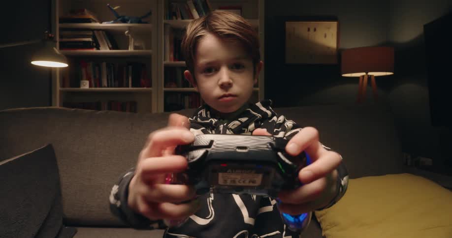 A young boy sits on a sofa concentrating on a video game before suddenly throwing his arms up and kicking his legs in the air to celebrate a victory in his dimly lit living room