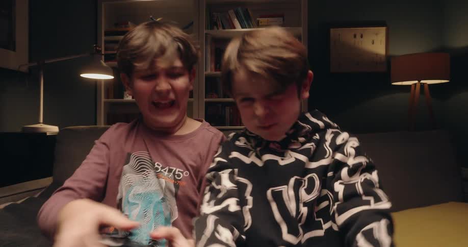 Two boys sit on a couch playing video games one boy smacking a game controller in a dimly lit living room looking forward with an excited and joyful mood