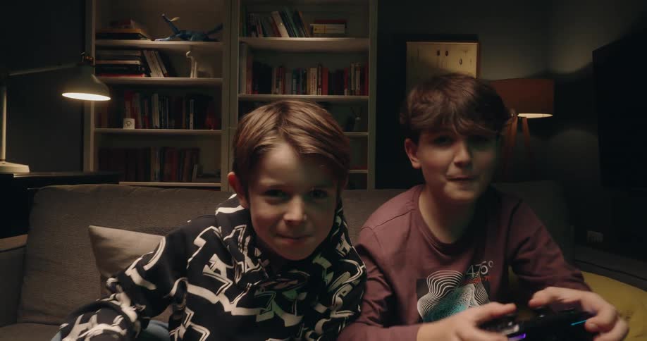 Two young boys sit on a grey sofa excitedly playing video games with controllers in hand while reacting to the action on screen in a dimly lit living room filled with books and home decor