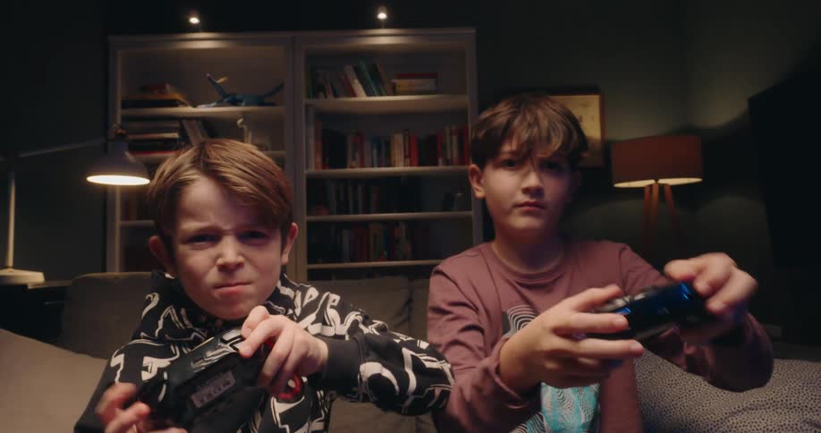 Two young boys sit on a couch in a dimly lit room focusing intently on a video game with determined expressions they grip their controllers tightly and lean forward surrounded by bookshelves and warm lamplight as they play