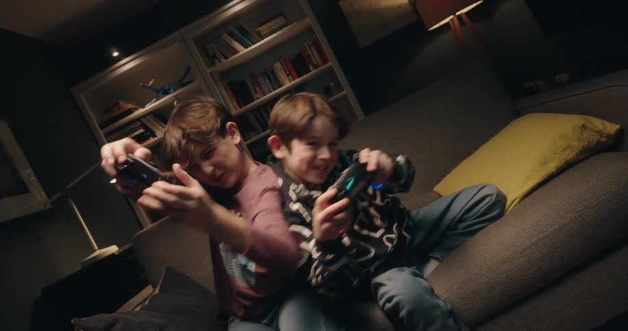 Two boys sit on a couch playing video games in a dimly lit living room looking forward with an excited and joyful mood