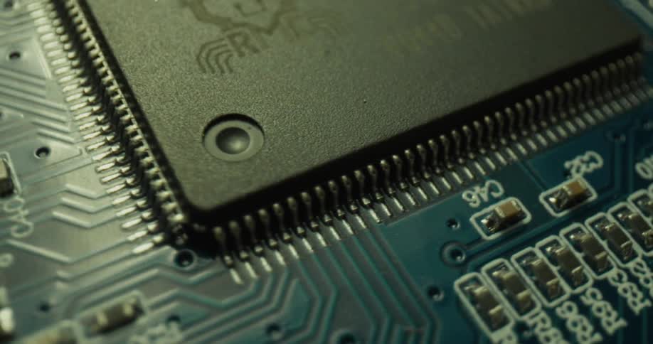 A closeup view shows the intricate details of a black microchip on a blue circuit board while light colors change and the microchip orbits
