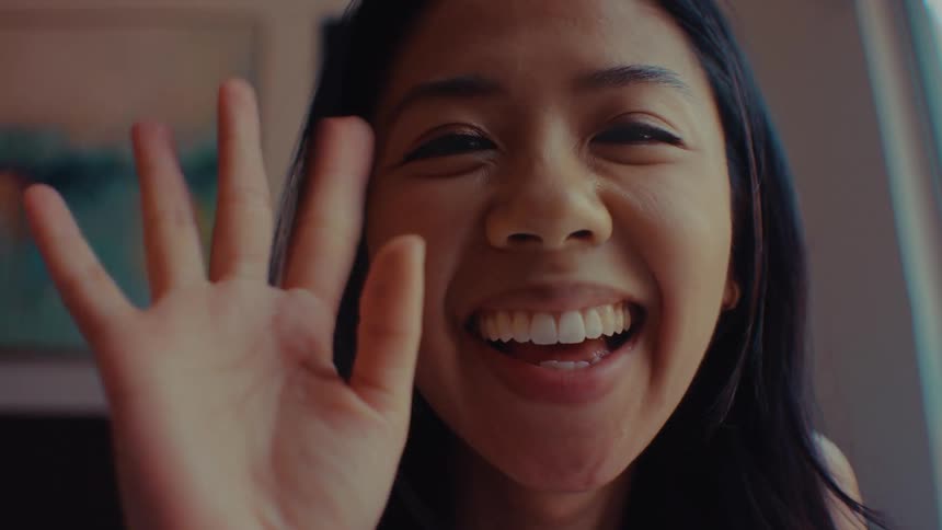 A young woman with long dark hair smiles brightly while looking directly into the camera lensshe raises her hand to give a cheerful wave appearing to greet someone during a video call