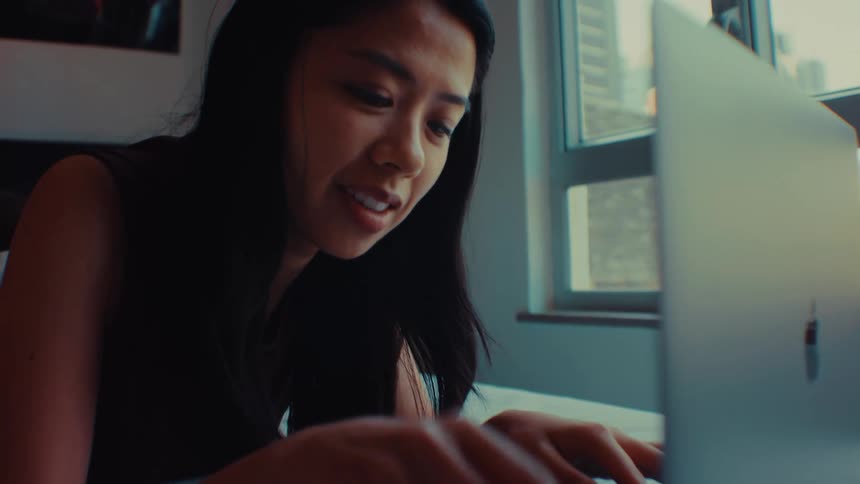 A young woman with long dark hair lies on a bed and works on a laptop in a sunlit room