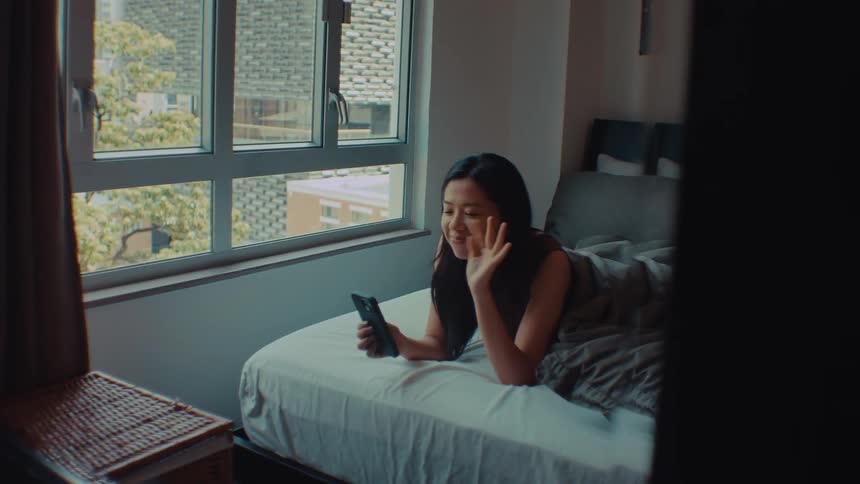 Young asian adult woman lying down in bed while smiling and waving at a smartphone indoors during day time
