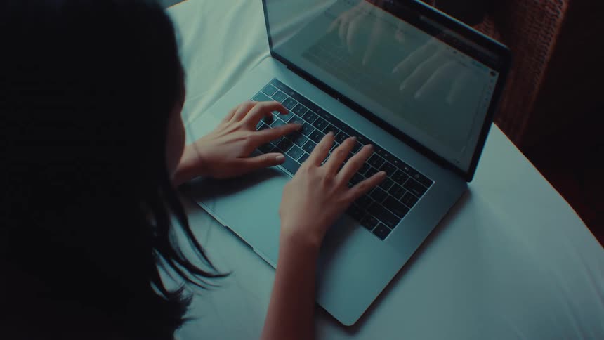 A female shown from the shoulders to head is sitting at a table typing on a laptop in a dimly lit room