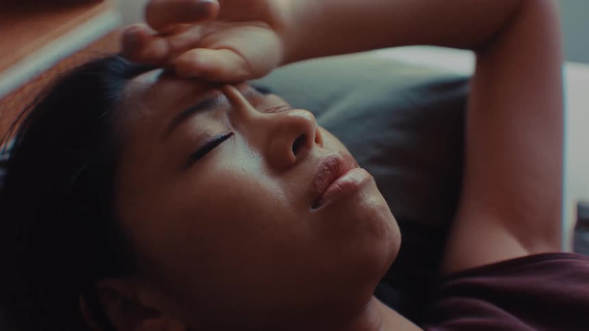 A woman lies down and rests a hand on her forehead sad and looks like she cried while looking upward in a dimly lit room