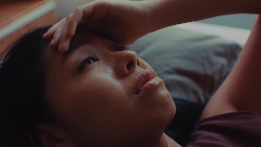 A woman lies down and rests a hand on her forehead sad and looks like she cried while looking upward in a dimly lit room